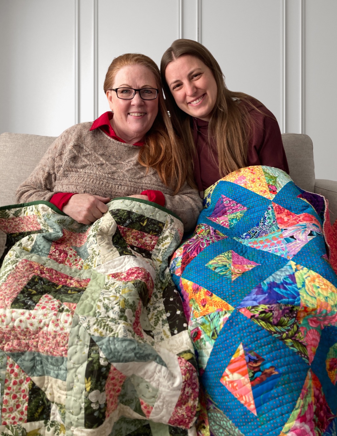 Cheryl and Marsali sitting together on a couch, each holding a quilt